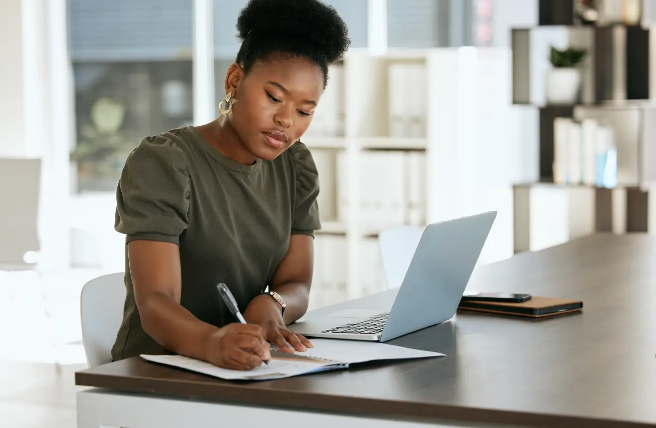 A person making notes while sitting at a desk with their laptop out